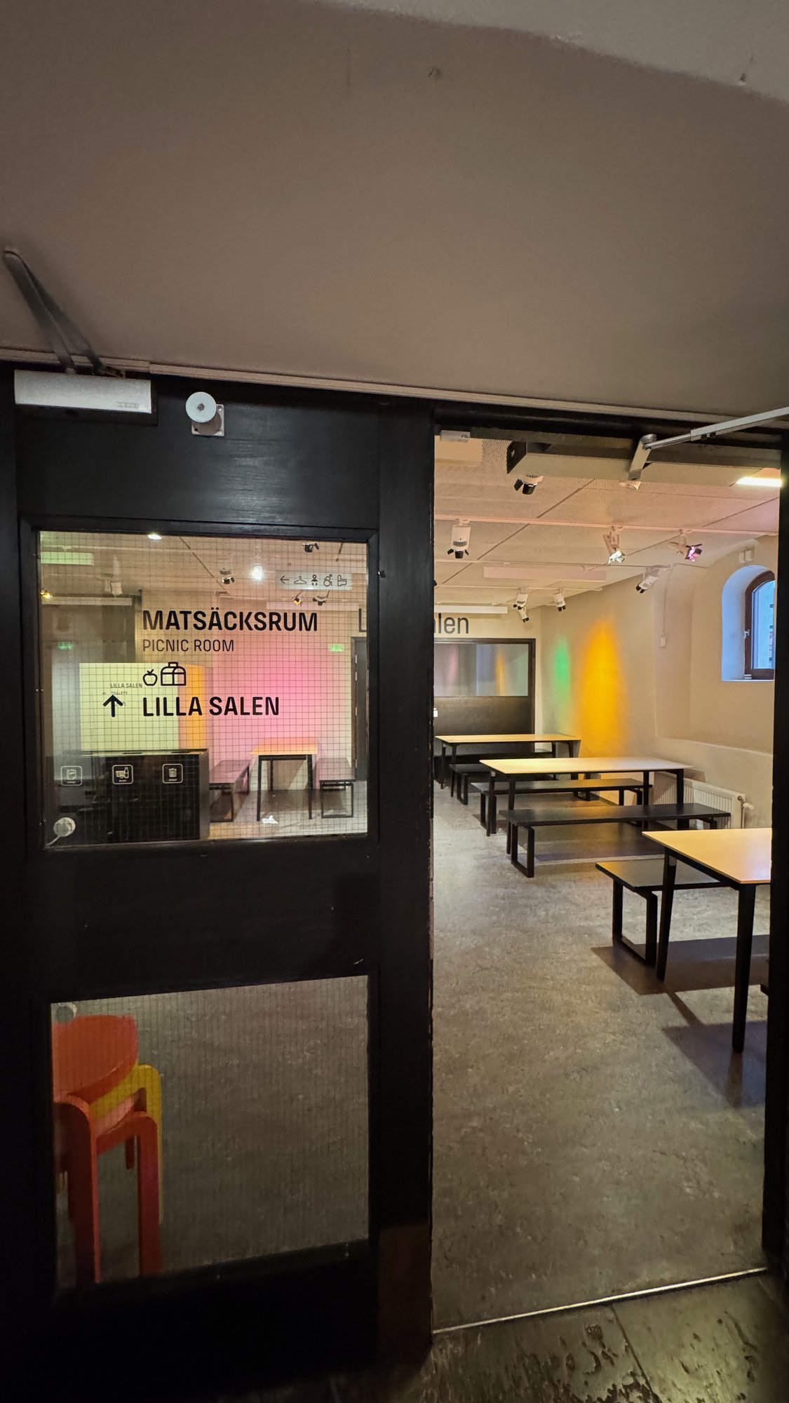 A picnic room with tables and benches behind a glass door with the sign Matsäcksrum at Scenkonstmuseet.