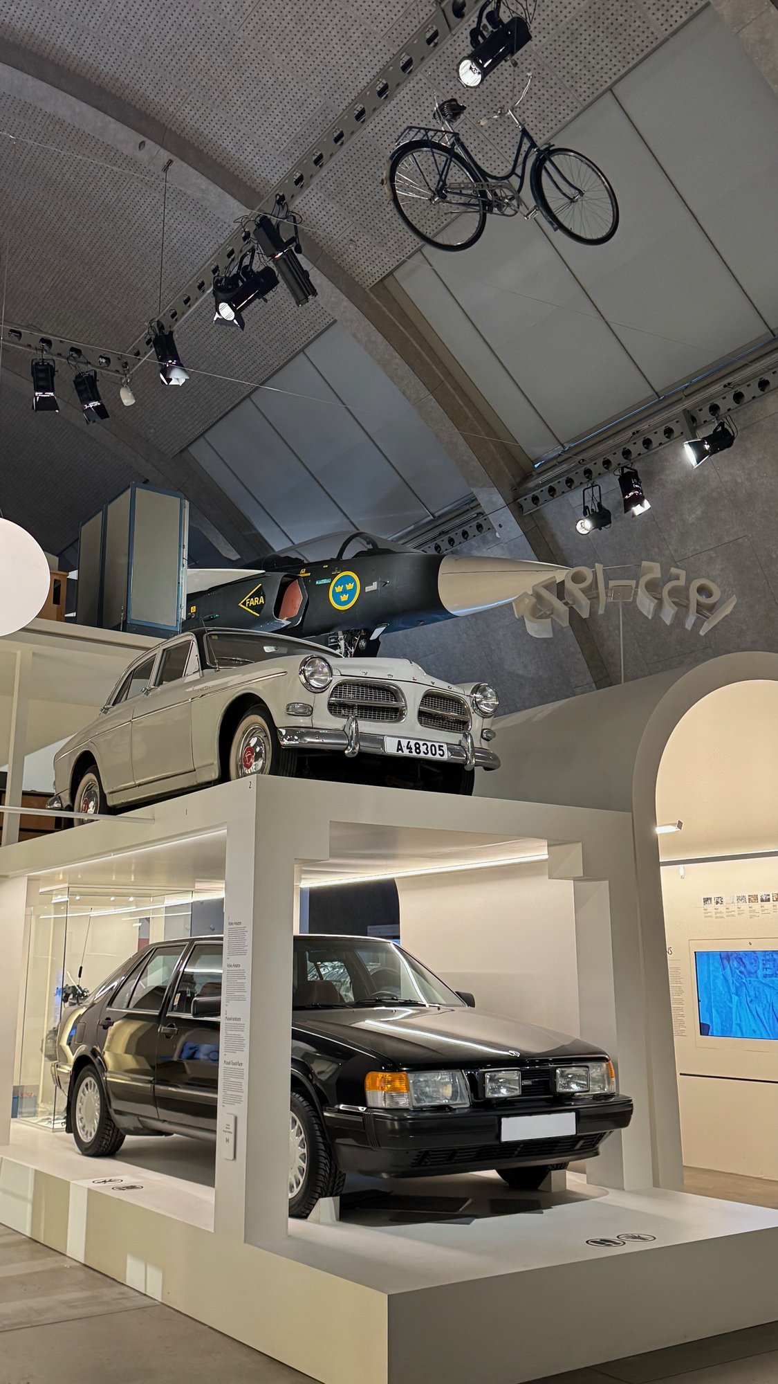 A black car in a glass box, a white car on top and an aircraft under the arched roof at Tekniska museet.