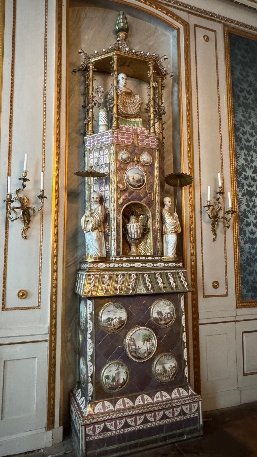 A tall tiled stove in chinoiserie style with painted medallions, small figures and a pagoda-like top, set in a white and gold panelled wall.