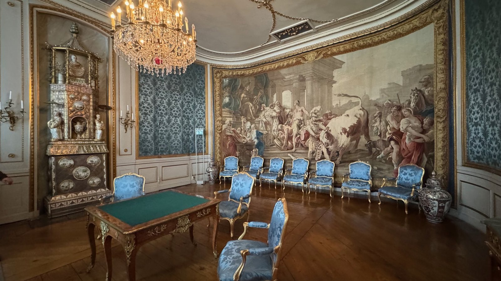 A drawing room with blue damask walls, blue upholstered chairs along the wall, a green card table and a large woven tapestry.