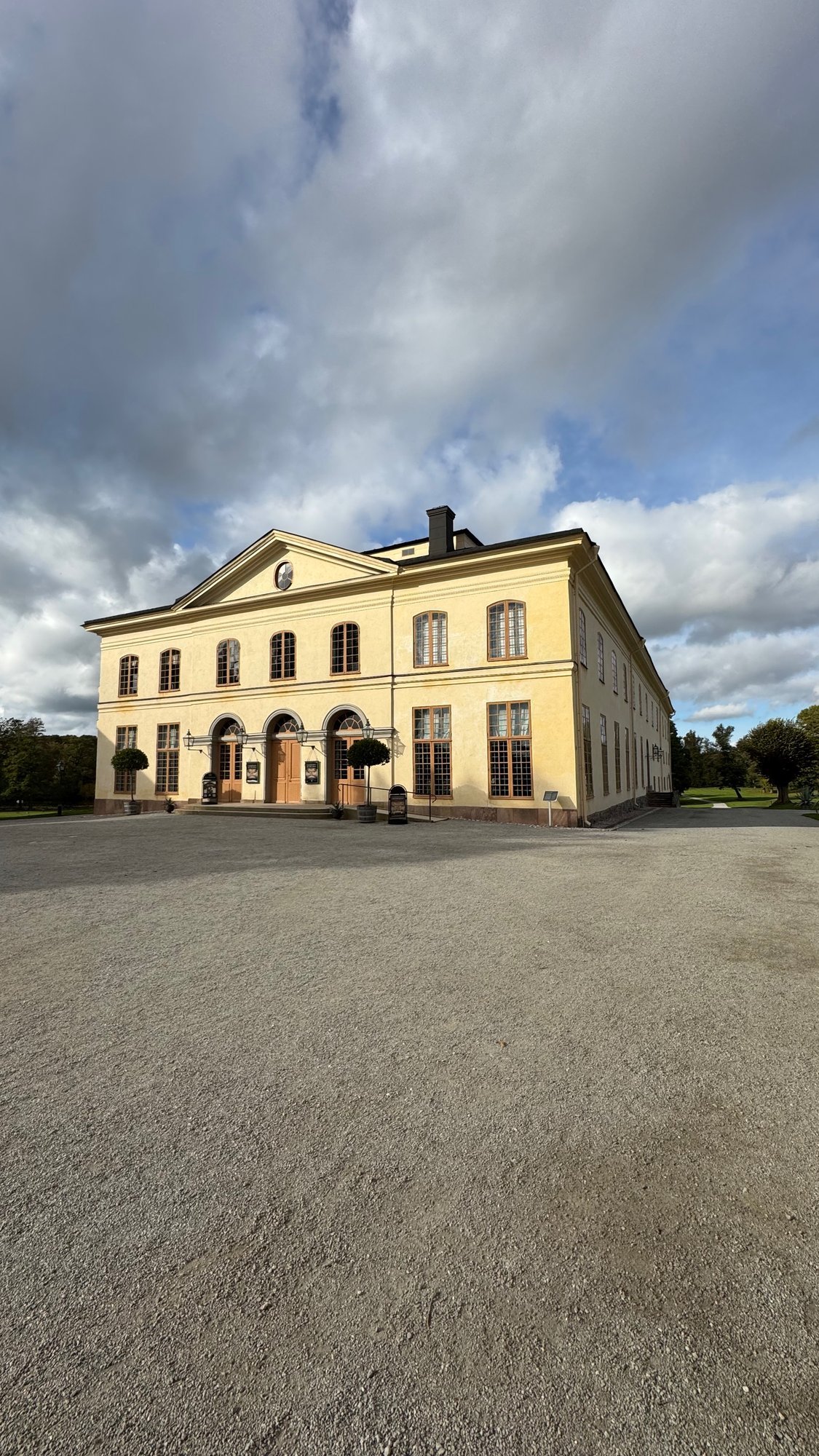 The pale yellow neoclassical building of the Drottningholm Court Theatre seen across a gravel yard under a cloudy sky.
