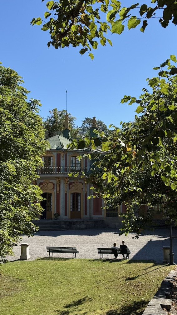 Two visitors resting on a garden bench at Drottningholm, with a pavilion visible among the trees behind them.