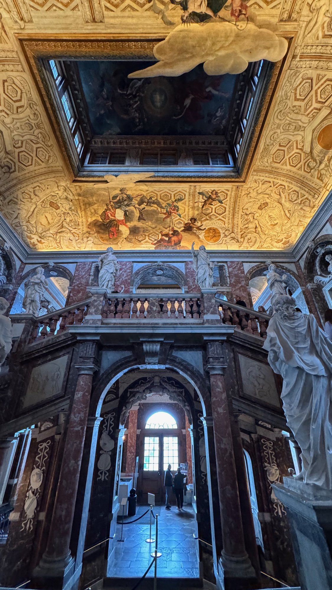 Looking up the grand staircase past marble statues toward a gilded and painted ceiling with a dark painted panel in the middle.