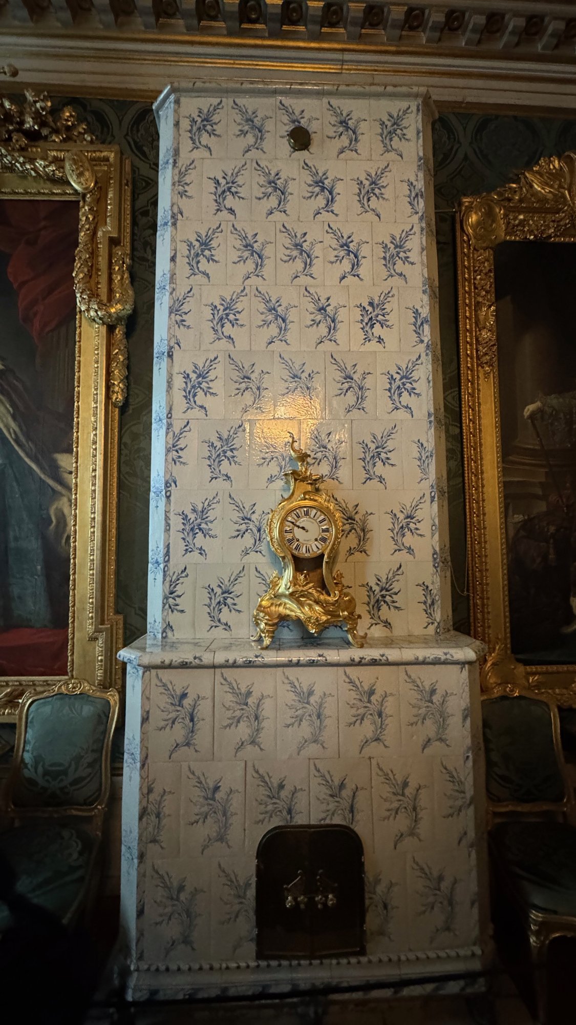 A tall white tiled stove with blue floral pattern and a gilded clock on top, between portraits in gilded frames in the Green Drawing Room.