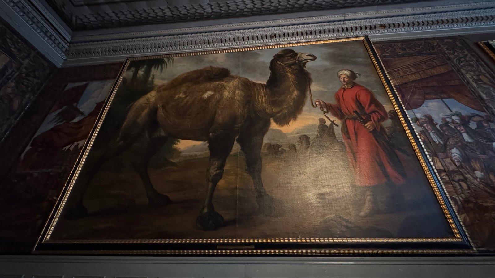 Large painting of a shaggy brown camel led by a man in a red coat and white turban, in a gilded frame on a gilt-leather wall.
