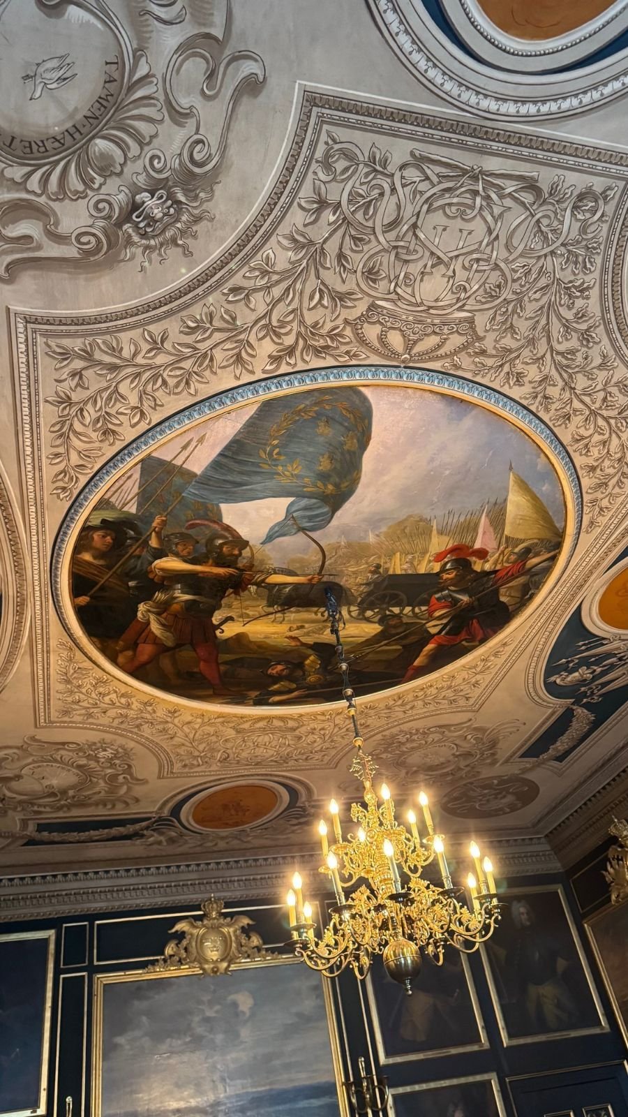 Ceiling of the Hall of Generals with white stucco, a painted oval of a battle and a gold chandelier above dark blue walls hung with portraits.