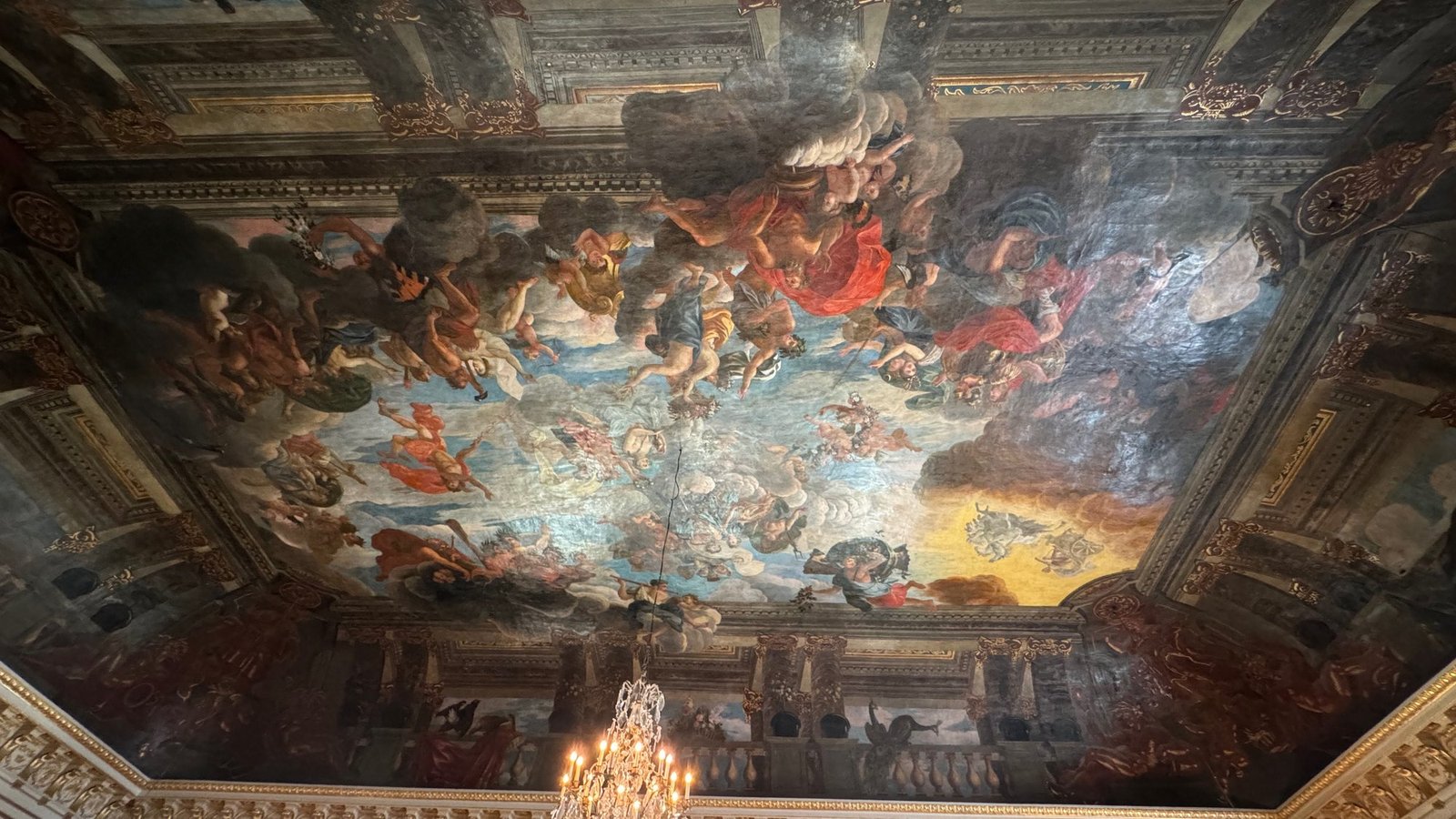 Painted ceiling with many figures in a gilded frame, with a crystal chandelier hanging below it.