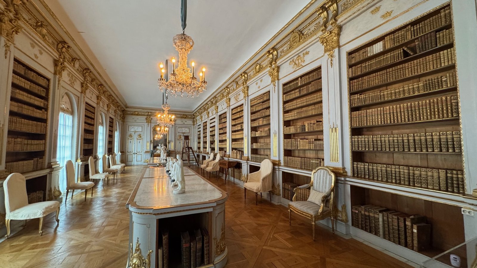 A long white and gold library room with tall bookcases on both sides, a table with busts down the middle and a chandelier.