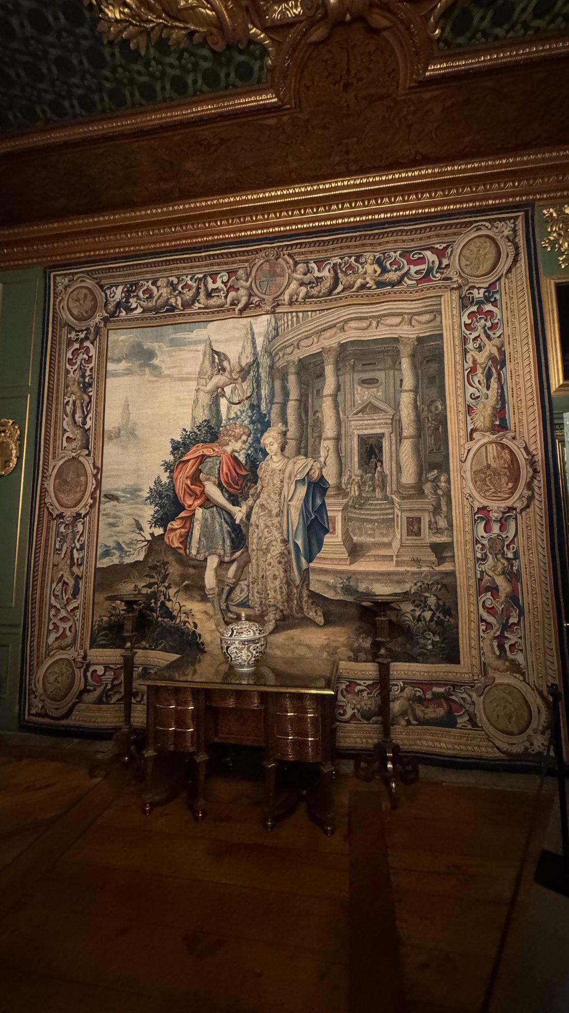 A woven tapestry with a decorated border showing two figures before a classical building, with a small table in front of it.