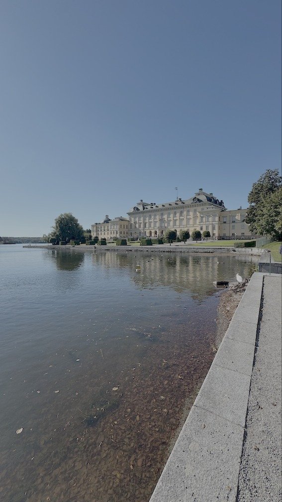 Drottningholm Palace seen across the still water of Lake Mälaren, its long baroque facade mirrored on the surface.