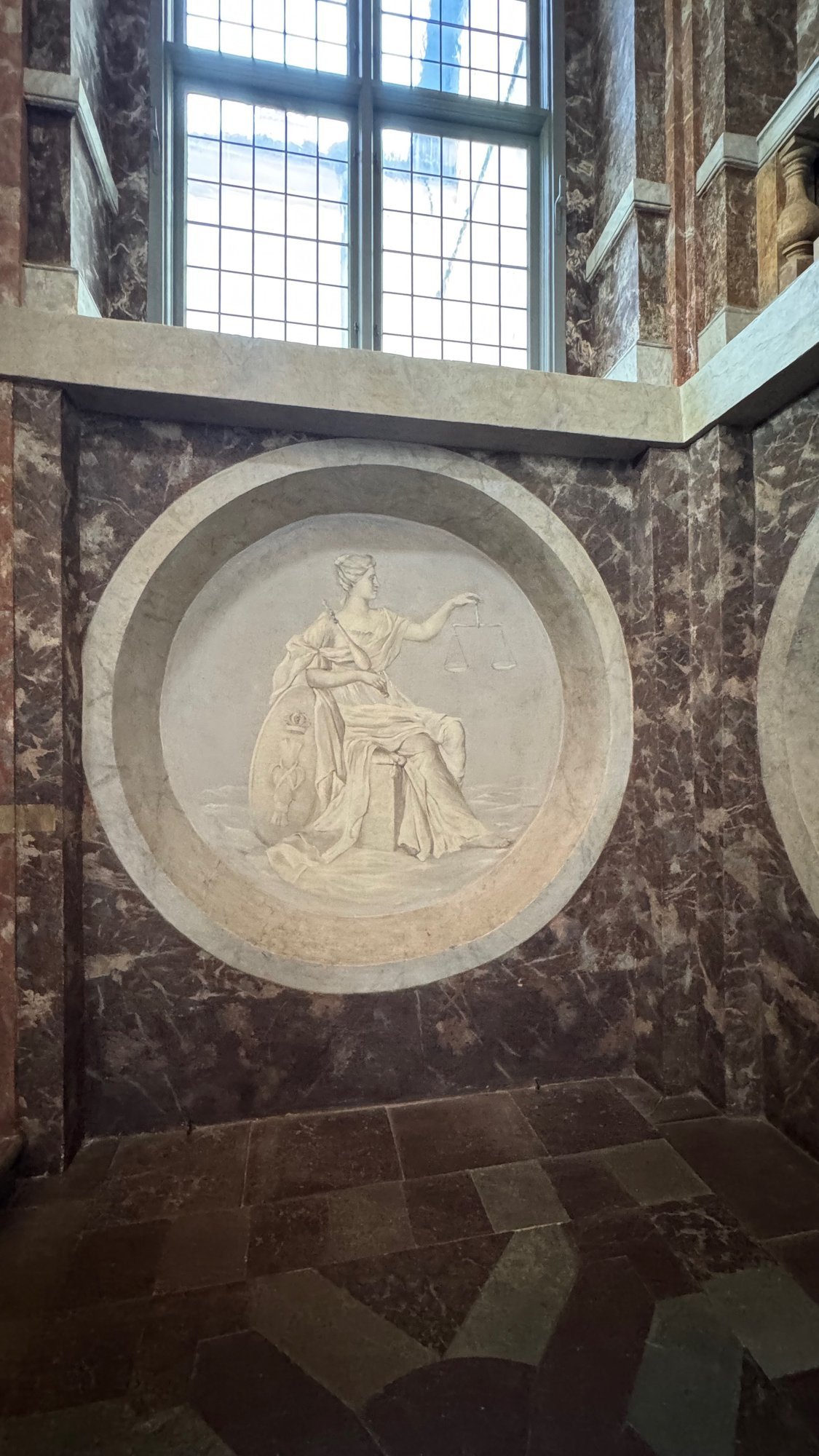 A round painted relief of Justice holding scales, set in a wall painted to look like brown and pink marble, under a leaded window.
