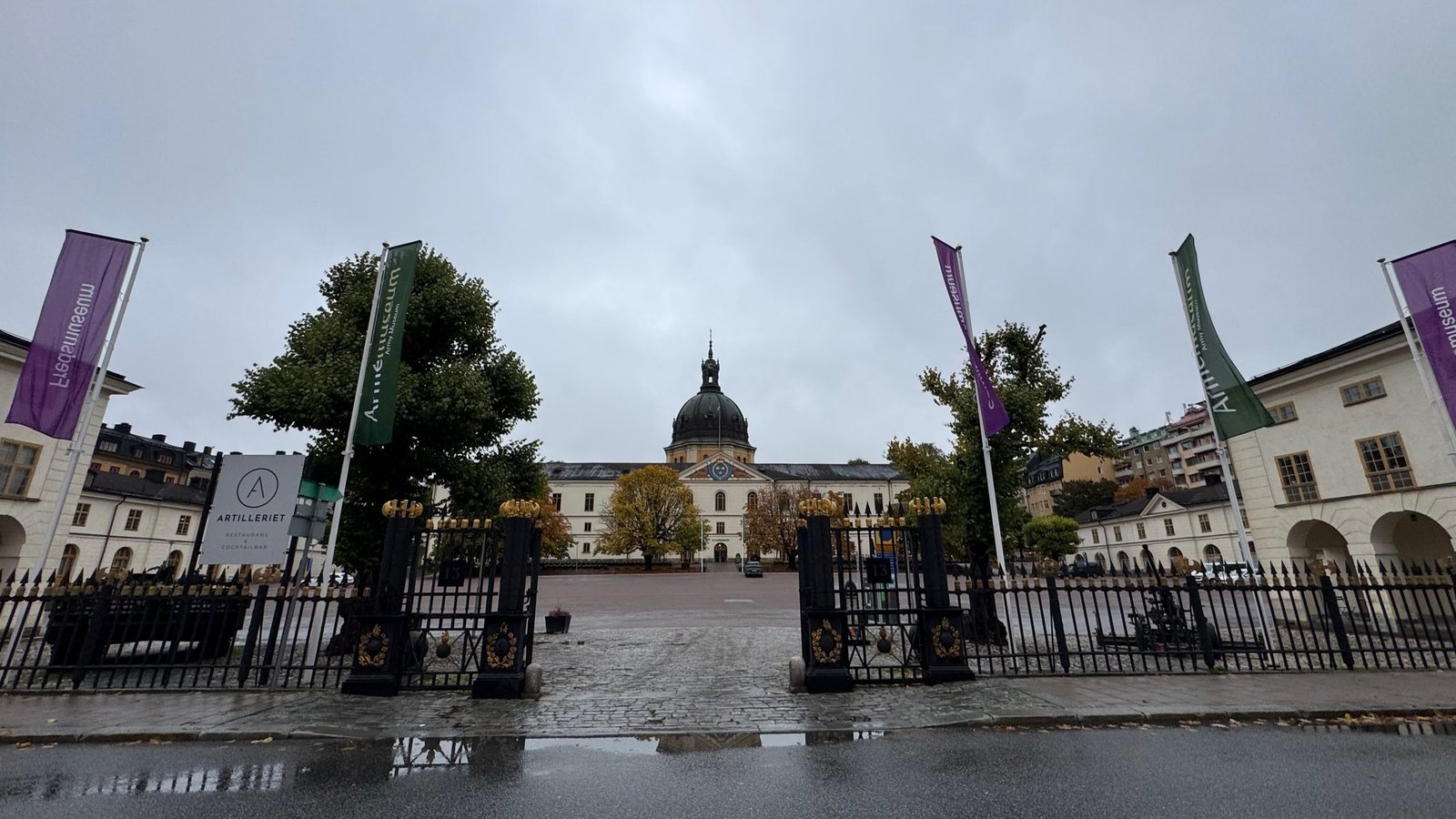 The Artillerigården courtyard of Armémuseum seen through an iron gate, with flags and a domed building.