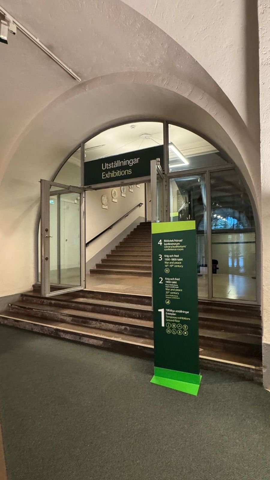 Stone stairs under a white arch lead to the exhibitions, with a green floor guide sign, at Armémuseum.