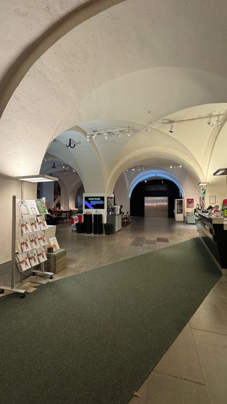 The reception area of Armémuseum with a green carpet, a brochure stand and a shop counter under vaults.