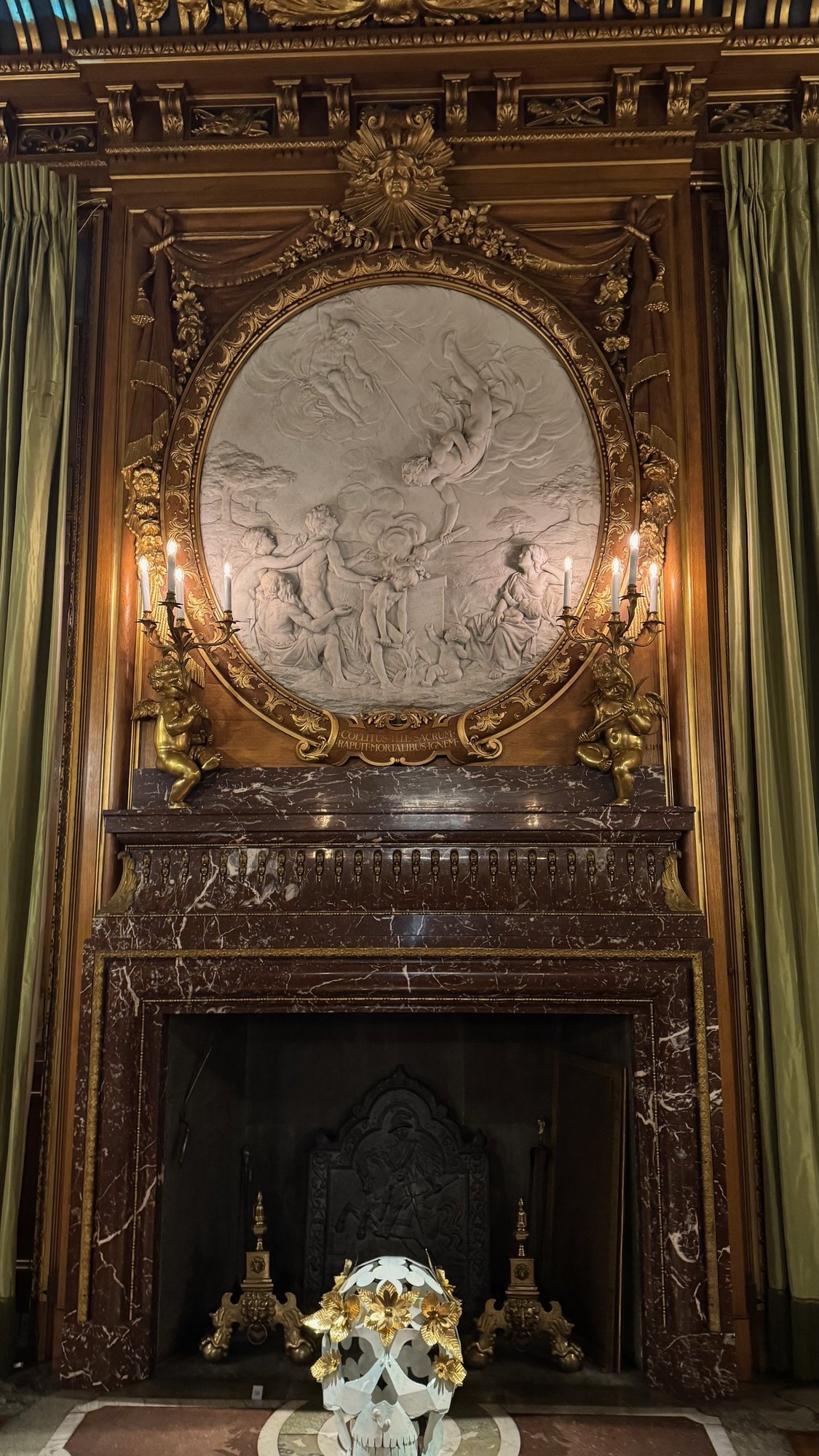 A marble fireplace with gold candle holders, a round white relief above it and a craft skull with gold flowers in front of it at the Hallwyl Museum.