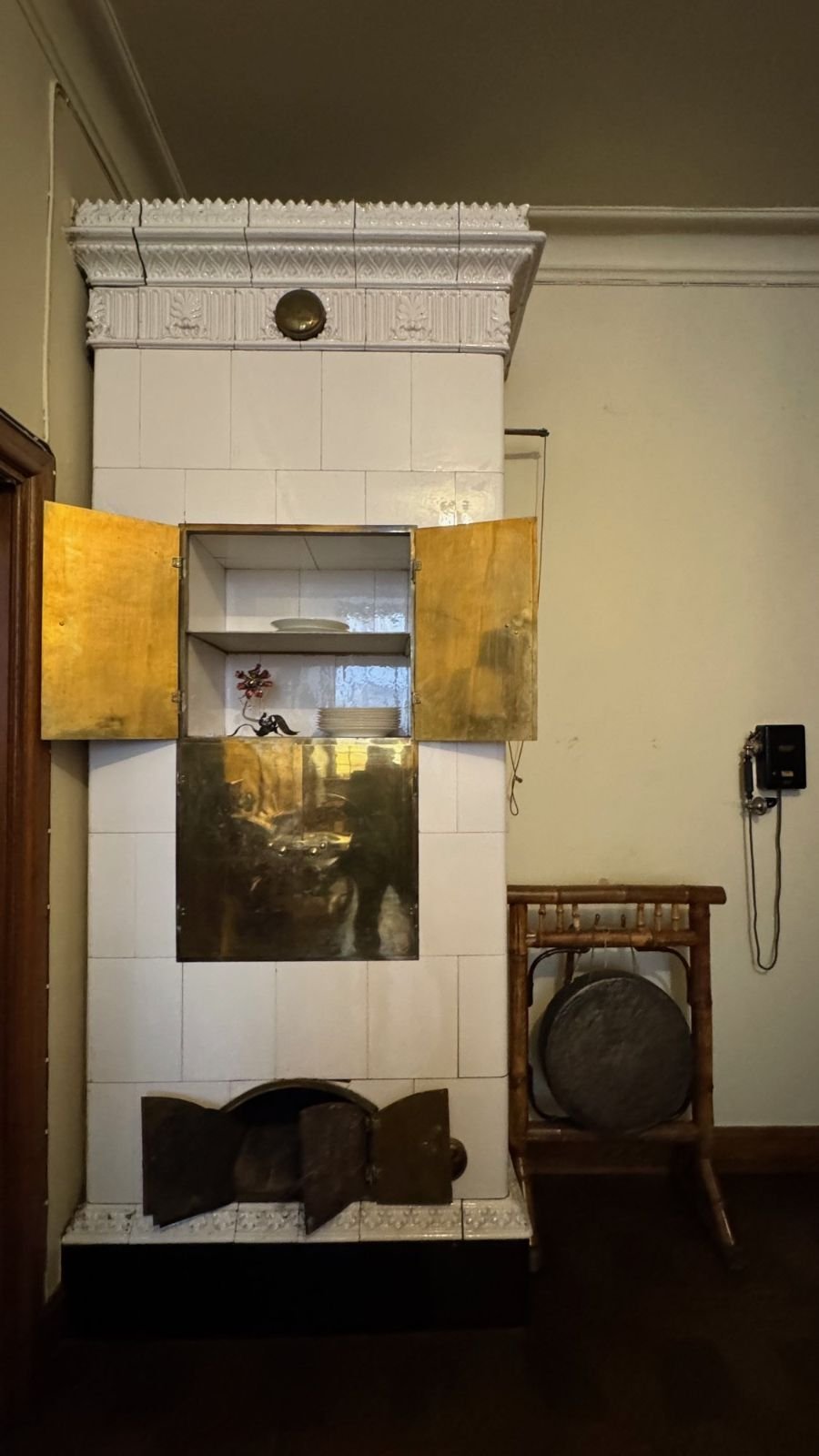 A white tiled stove with open gold doors next to a wall telephone and a gong at the Hallwyl Museum.