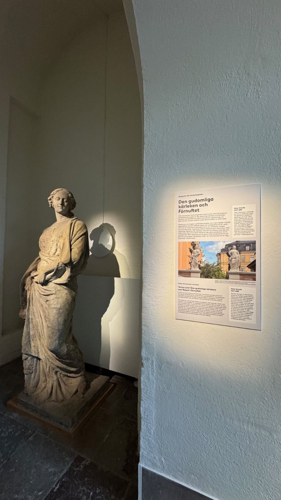 A stone statue of a woman stands in a lit niche next to an information panel at the Stockholm City Museum.