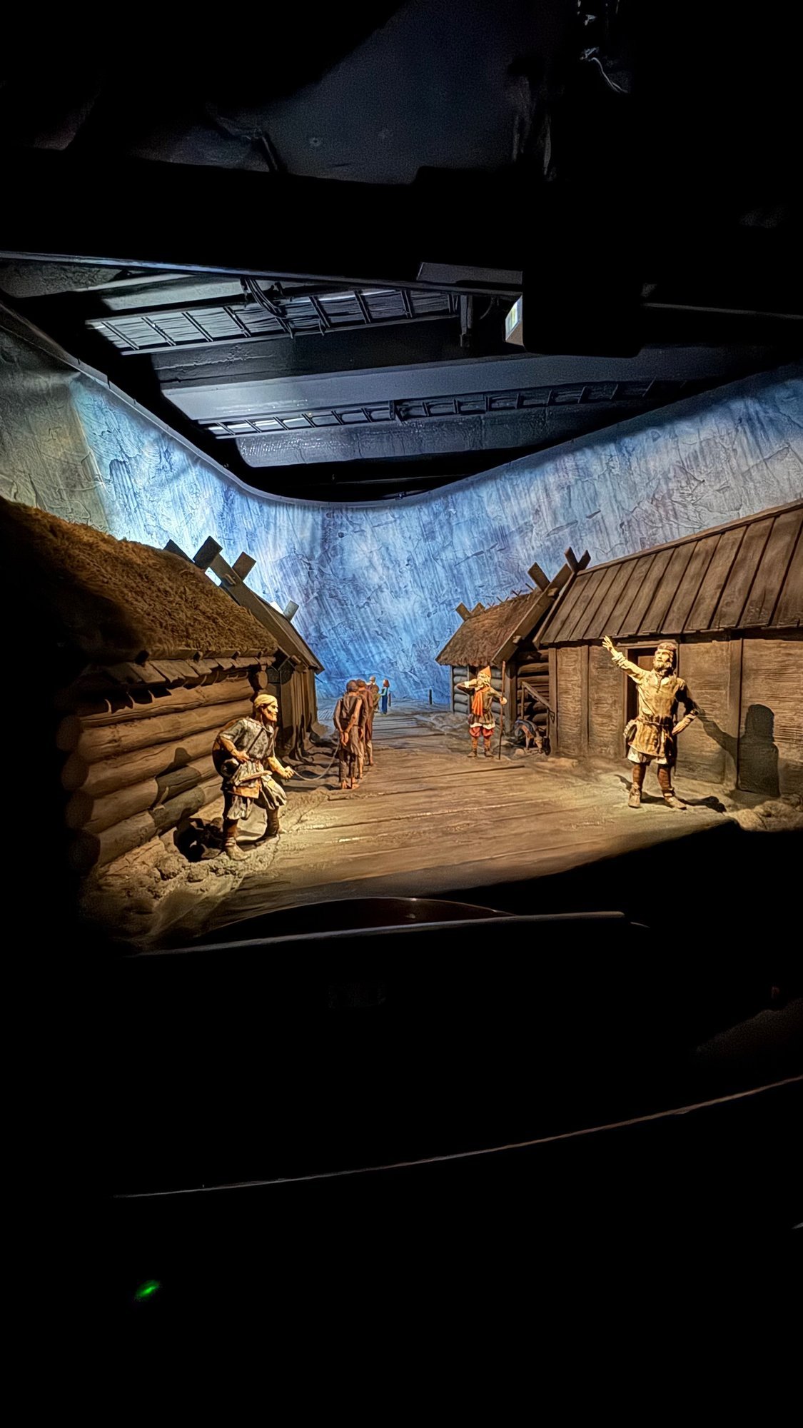 A lit model of a Viking village, with log houses and small figures in a lane, in a dark room.