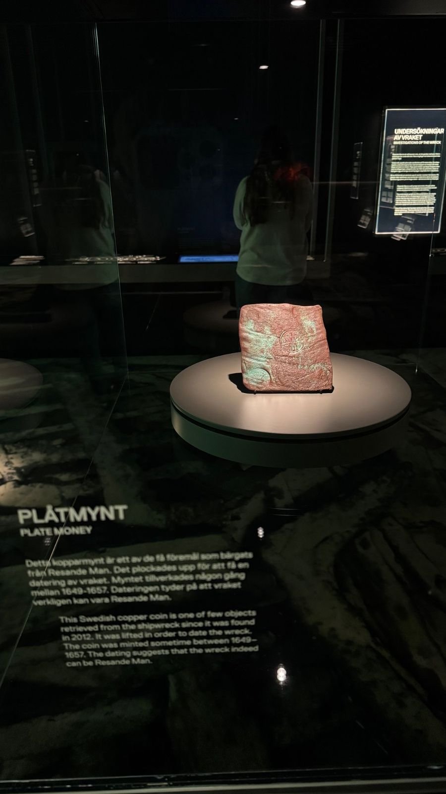 A square copper plate coin on a round stand in a dark glass case, found at the wreck of Resande Man, at Vrak Museum of Wrecks.