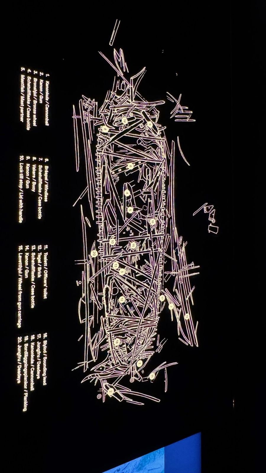 A white and purple drawing of the timbers of a shipwreck on a black wall, with a numbered list beside it, at Vrak Museum of Wrecks.