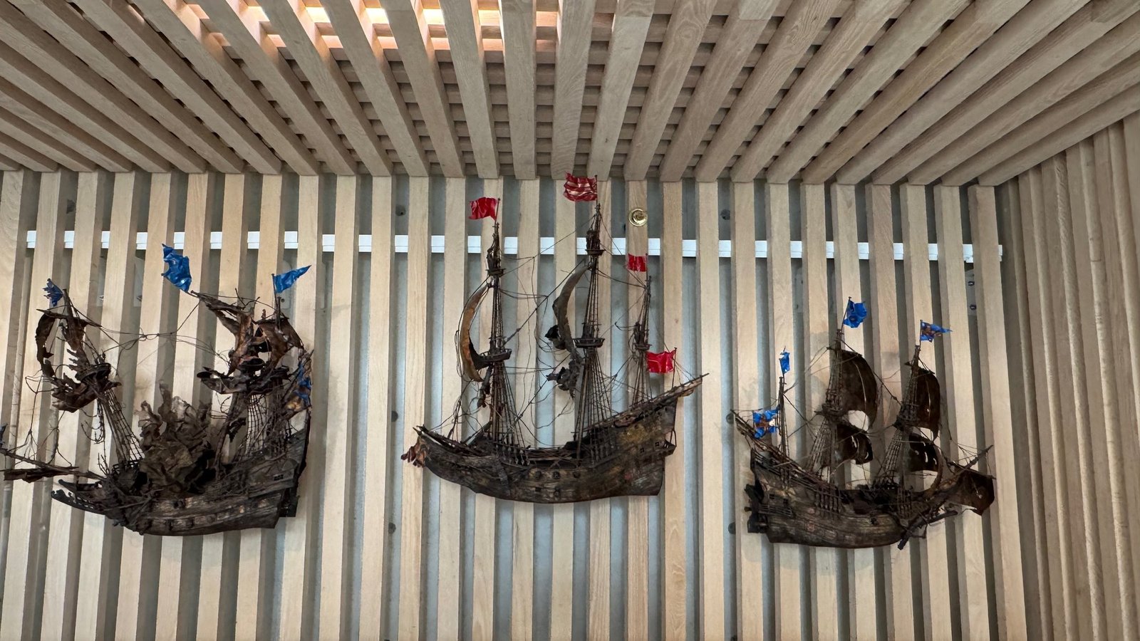 Three dark copper ship sculptures with torn sails and small red and blue flags, on a wall of light wooden slats at Vrak &ndash; Museum of Wrecks.