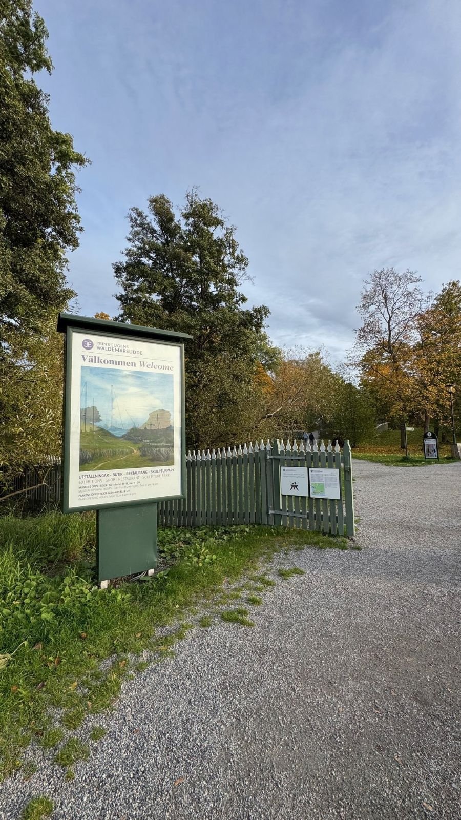 A welcome sign and a green wooden fence at the park entrance of Prince Eugen’s Waldemarsudde, with a gravel path.