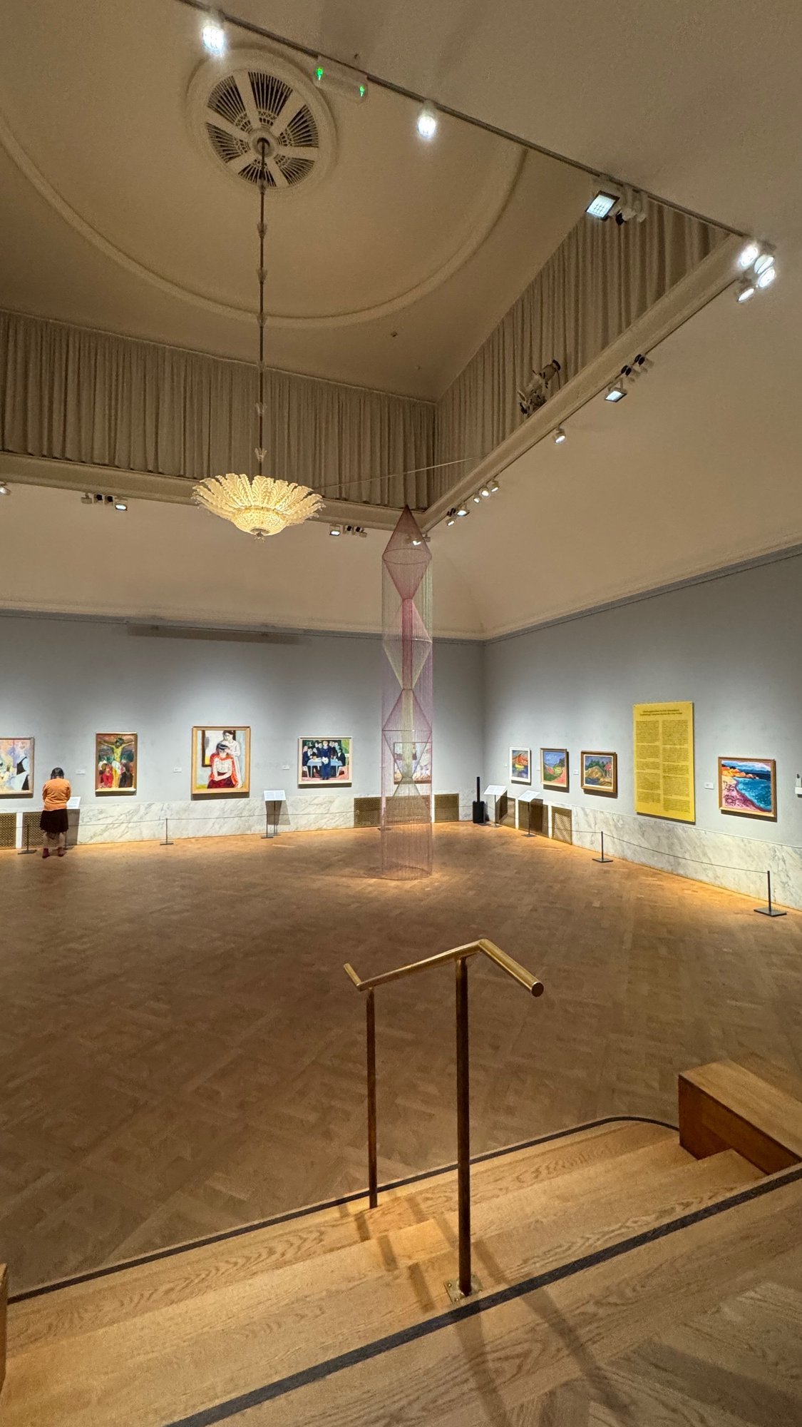 A large gallery hall with paintings, a hanging textile work and a crystal lamp, seen from the stairs at Waldemarsudde.