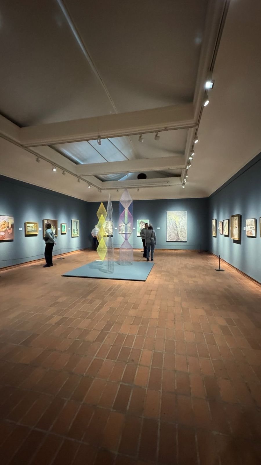 A gallery room at Waldemarsudde with paintings on blue walls, a brick floor and a colorful hanging installation.