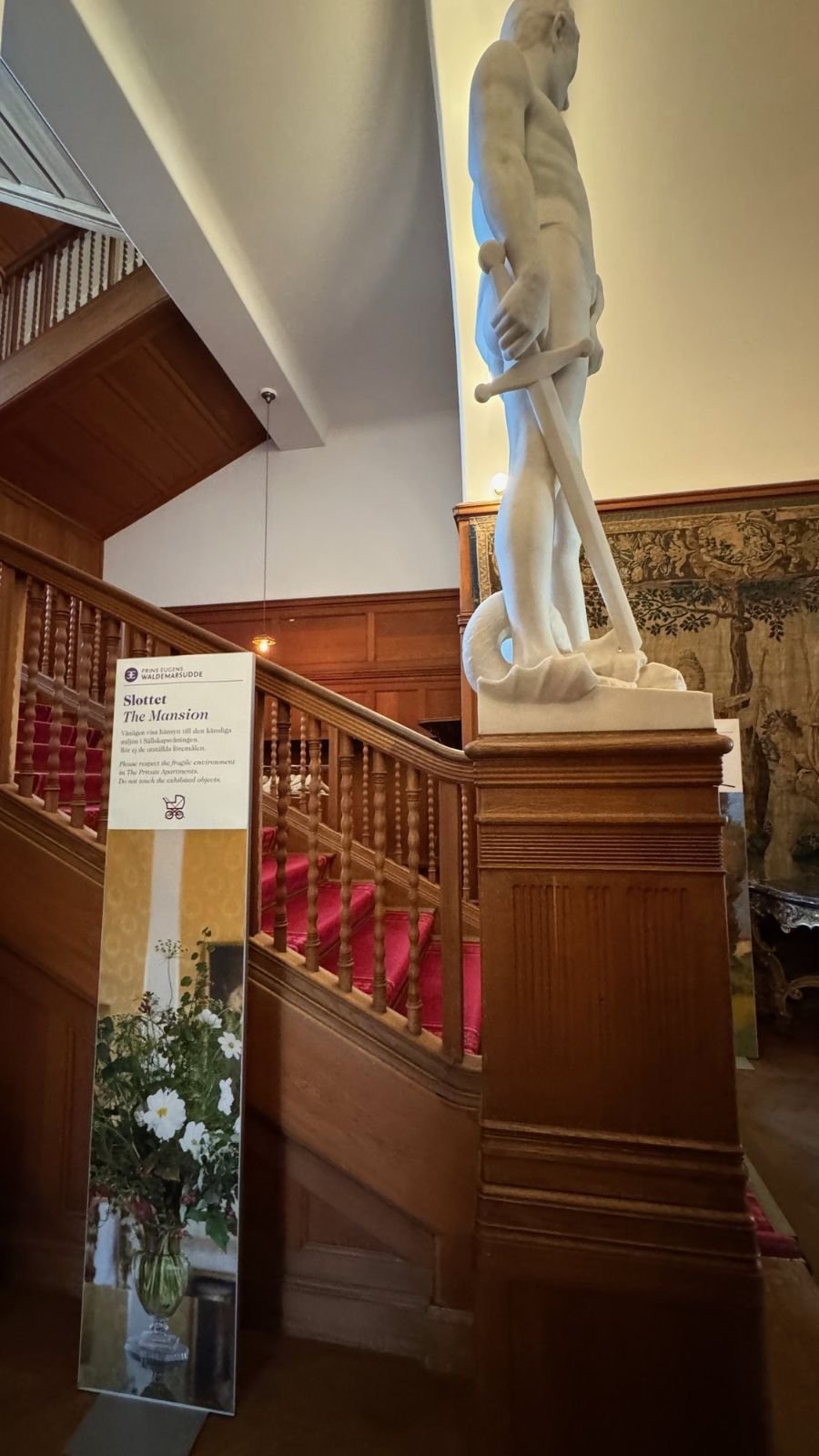 A white marble sculpture of a man on a wooden newel post by the red-carpet staircase in the Waldemarsudde mansion.