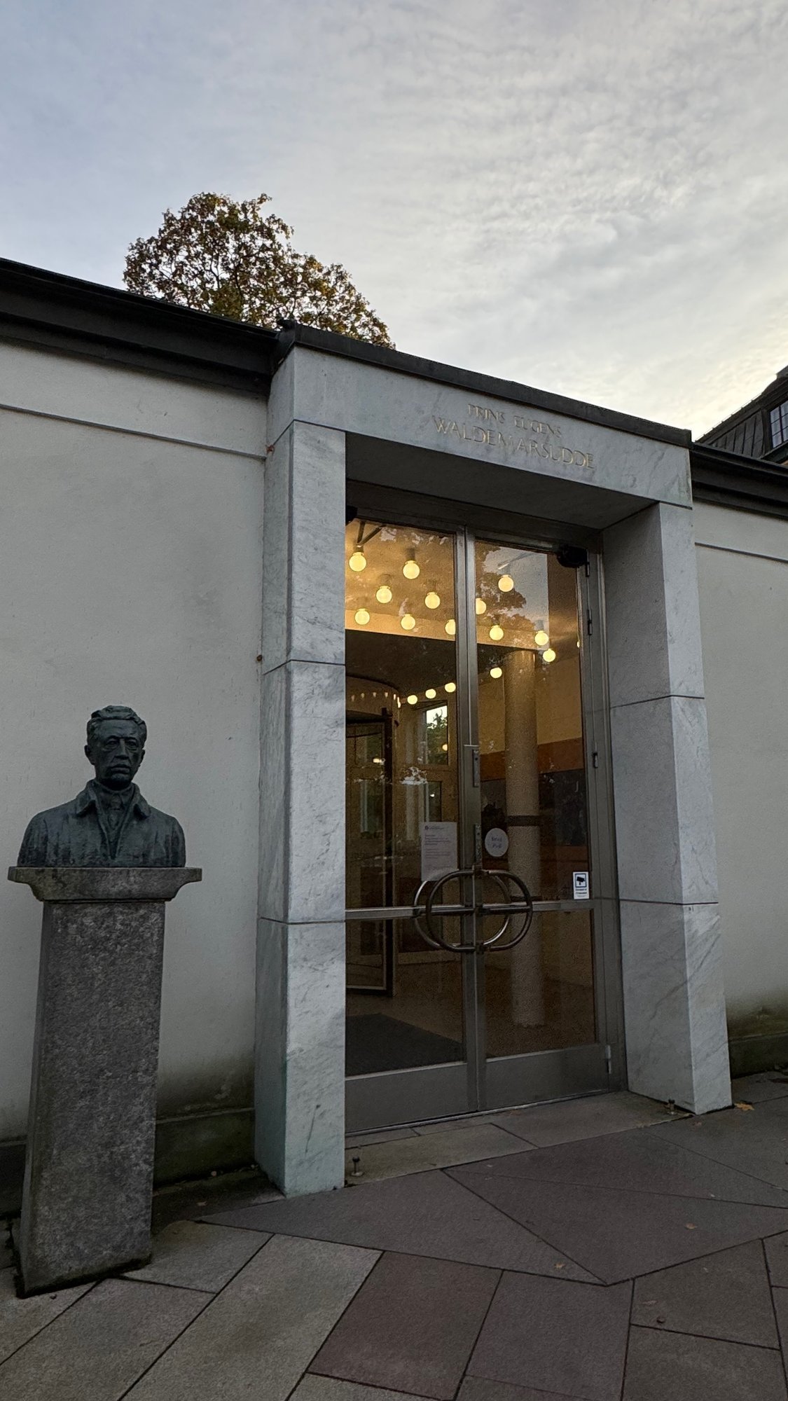 The glass entrance doors of the gallery building at Waldemarsudde with a bronze bust beside them and a flat stone path.