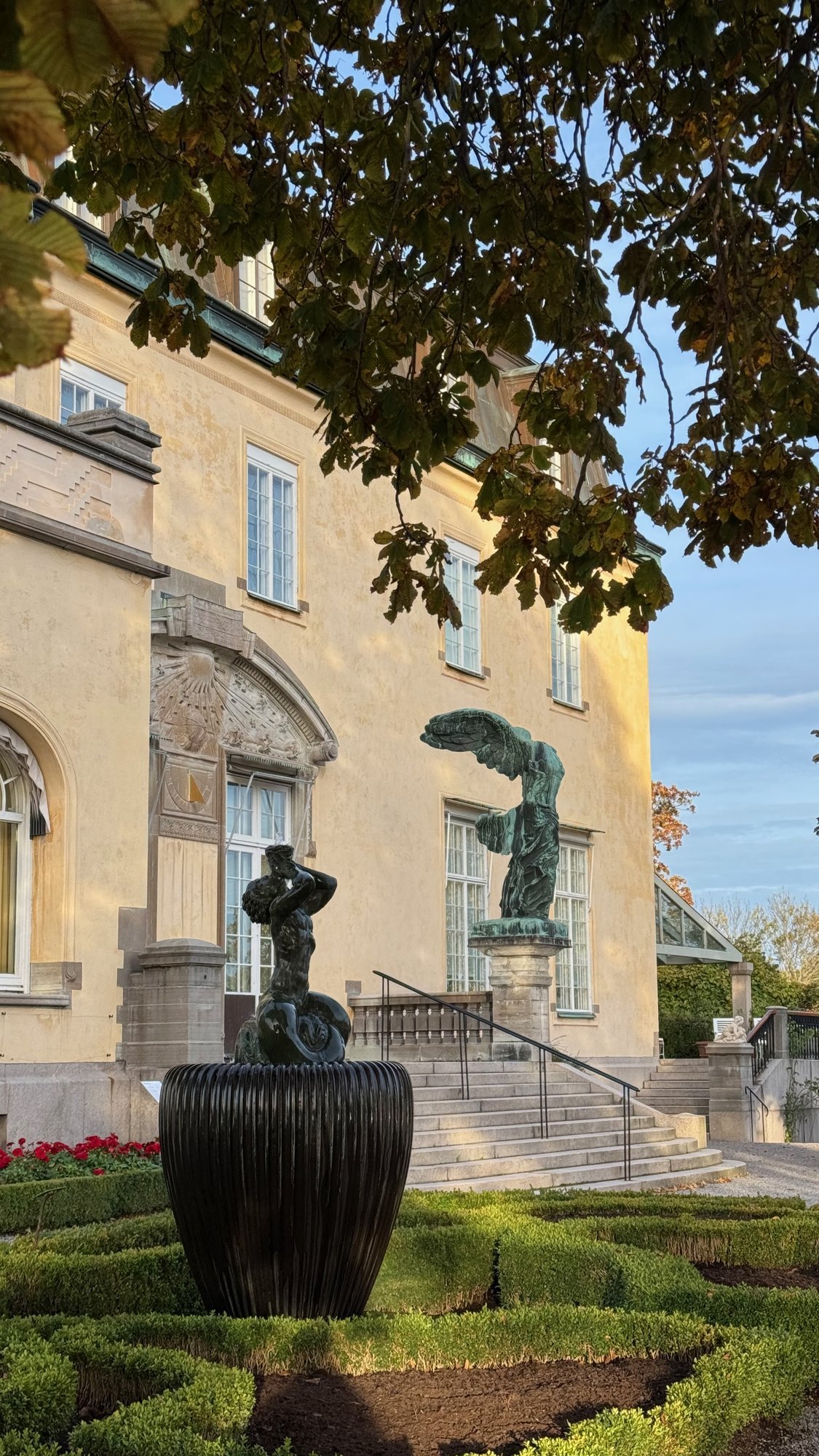 A bronze sculpture on a dark urn in a low hedge garden in front of the Waldemarsudde mansion.