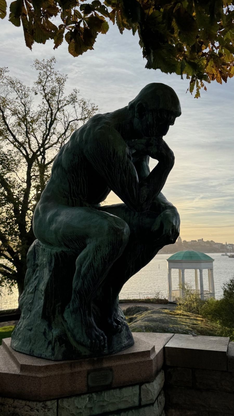 The Thinker by Rodin seen against the evening sky with the water and a white pavilion behind it at Waldemarsudde.