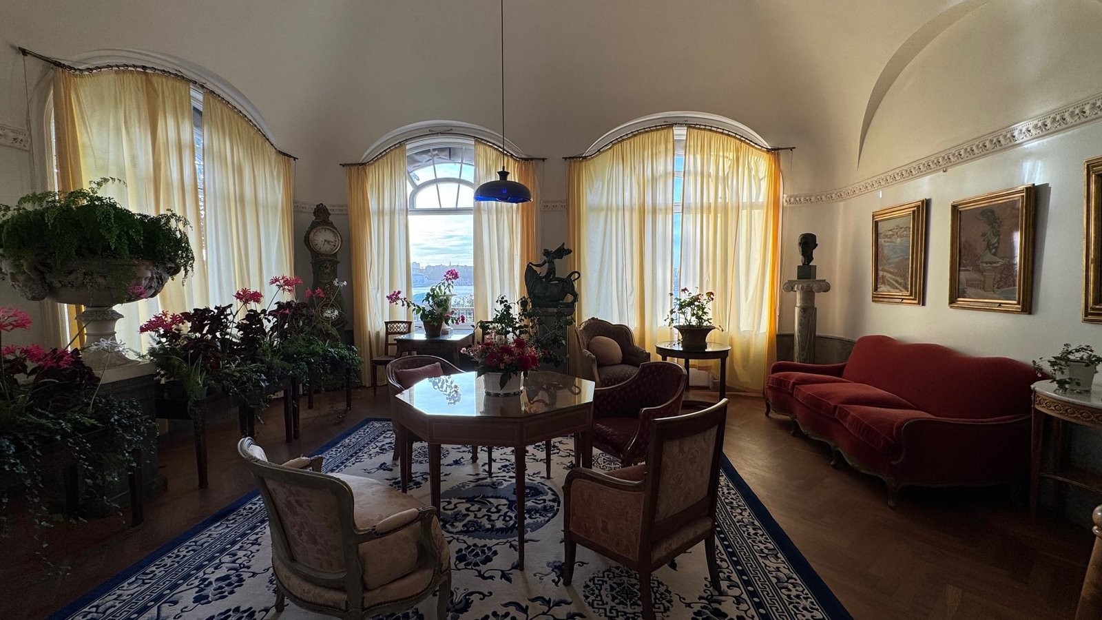 A room with arched bay windows, yellow curtains, plants, a red sofa and paintings in the Waldemarsudde mansion.