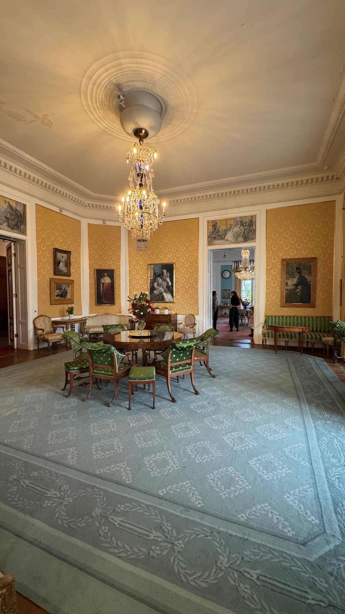 A drawing room with yellow walls, a crystal chandelier, green chairs and a large blue-grey carpet at Waldemarsudde.