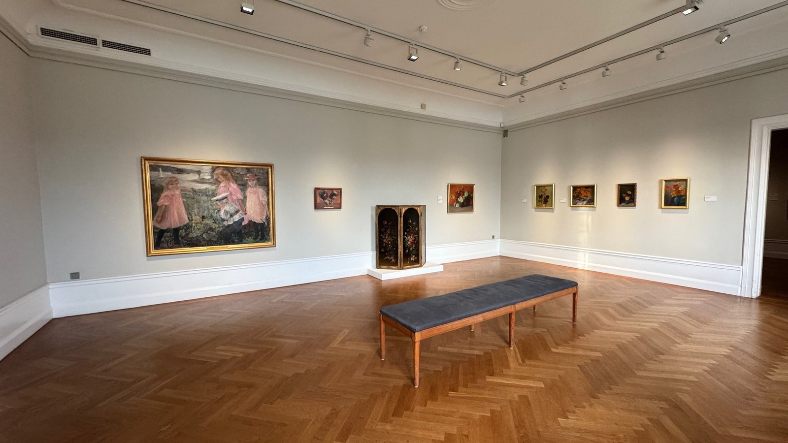 A gallery room with paintings on white walls, a herringbone floor and a long bench at Waldemarsudde.
