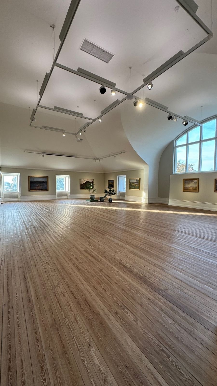 A large bright room with a wooden floor, paintings and arched windows at Waldemarsudde.