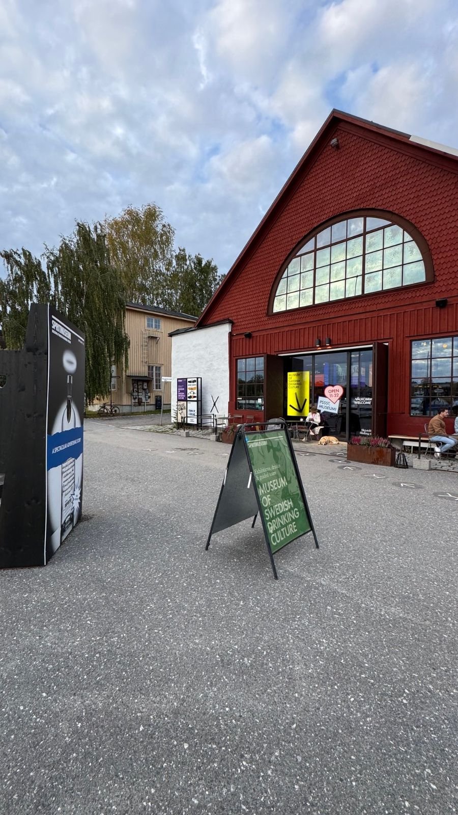 The red wooden building of Spritmuseum on Djurgården with a large arched window, open doors and a green sign that says Museum of Swedish Drinking Culture.