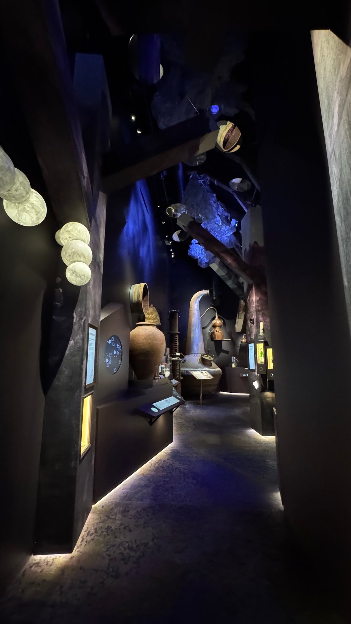 A dark exhibition corridor at Spritmuseum with blue light, old copper stills and glass cases under wooden roof beams.