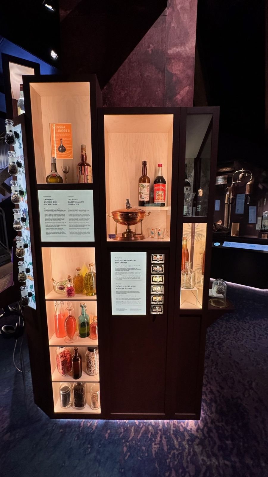 A tall lit cabinet with old bottles, a copper bowl and text labels in the dark exhibition at Spritmuseum.