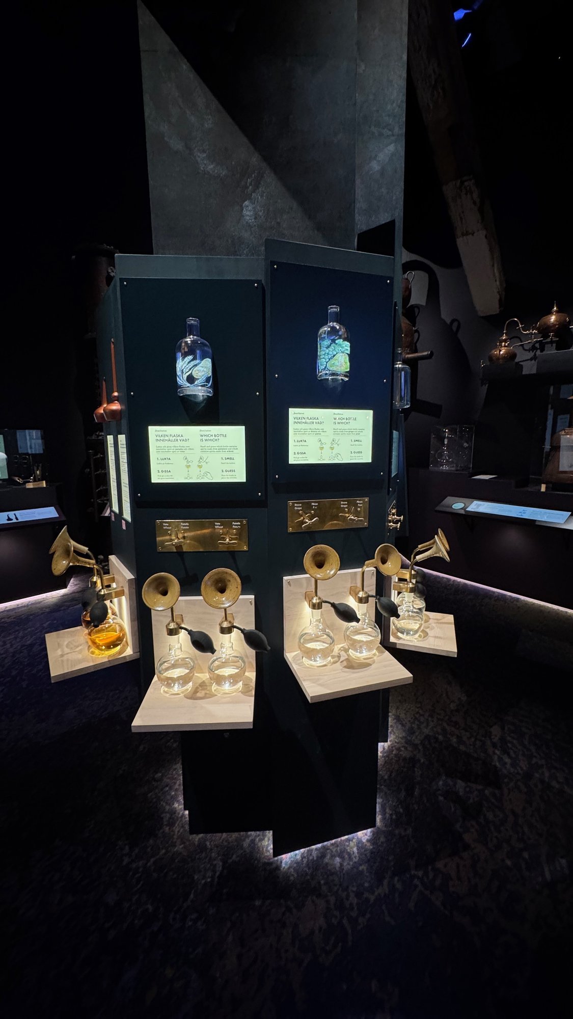 Brass horns over glass bottles on white tables, with two lit bottles on screens behind them, at Spritmuseum.