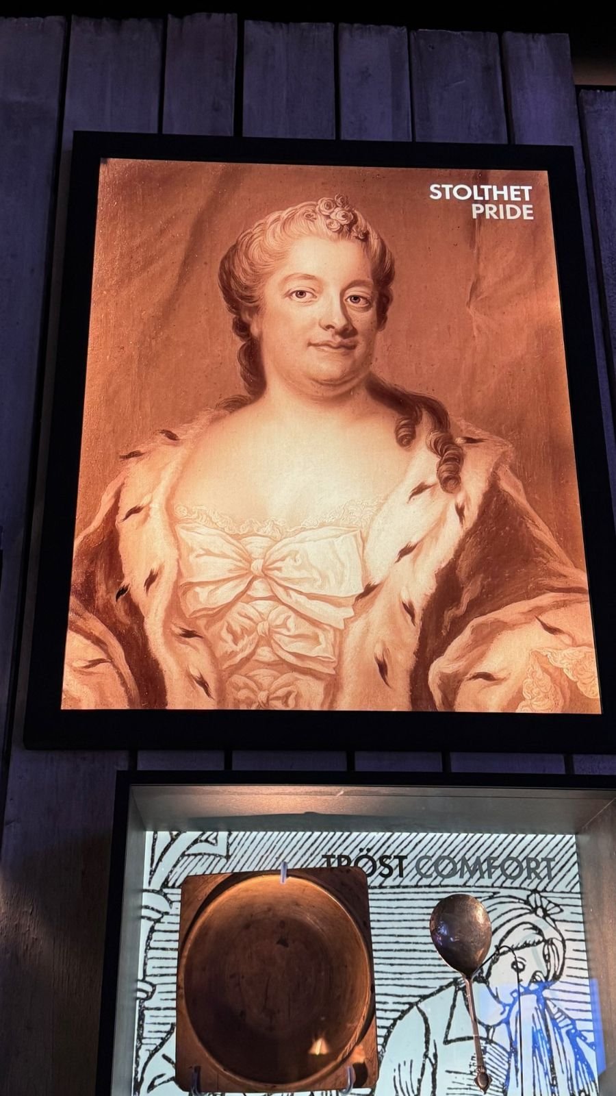 A large portrait of Eva De la Gardie hangs in the exhibition at Spritmuseum, with a display about comfort below it.