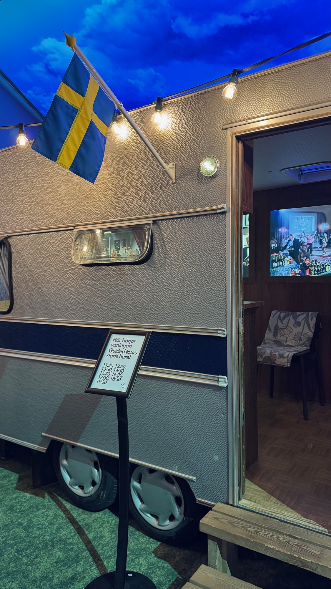 An old grey caravan with a Swedish flag and string lights in the exhibition at Spritmuseum.