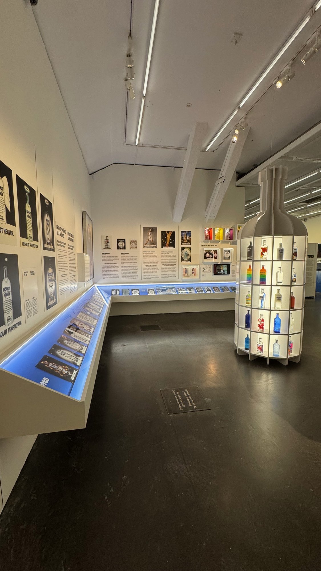 A bright exhibition room at Spritmuseum with Absolut ads on the walls, a long glass case and a bottle-shaped display.