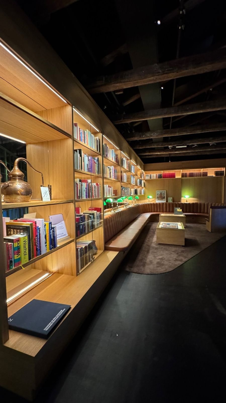 A library lounge at Spritmuseum with wooden bookshelves, a long sofa, green lamps and a copper pot.