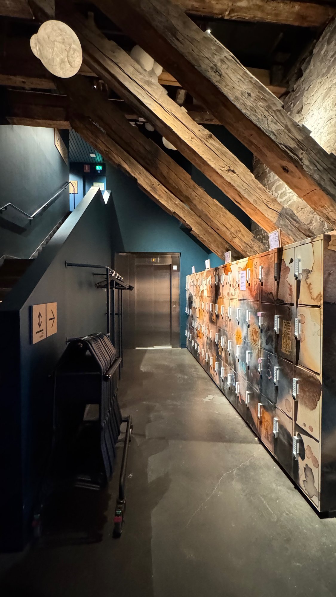 A lift door, coat racks and a wall of lockers by the stairs at Spritmuseum, with a sign for the lift.