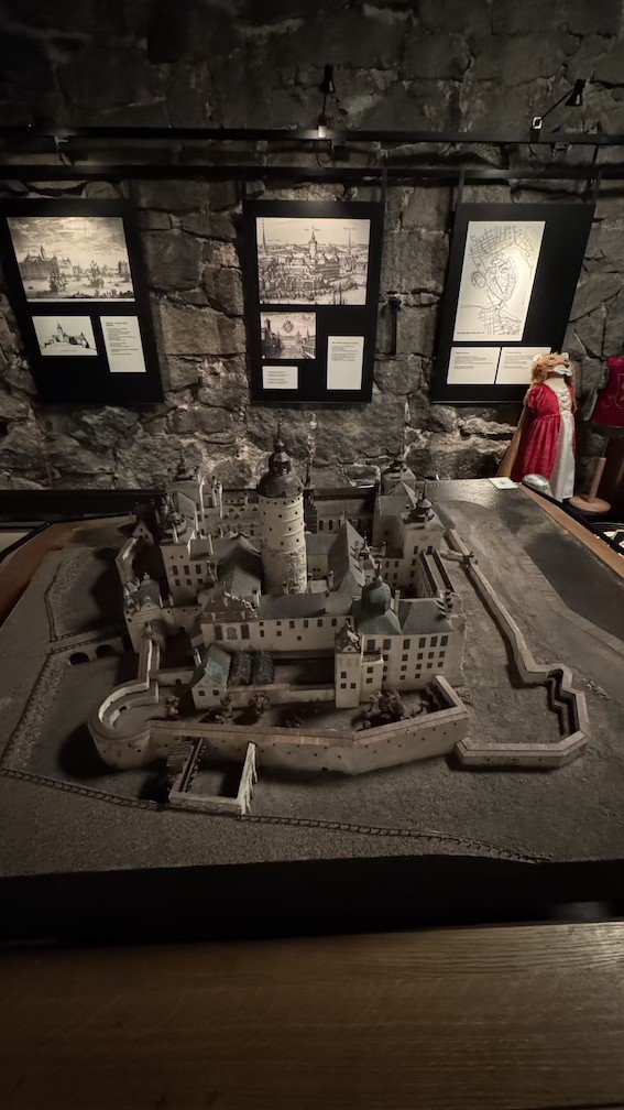 Detailed wooden scale model of the Tre Kronor Palace as it looked in 1663, shown on a display table with historical engravings mounted behind it.