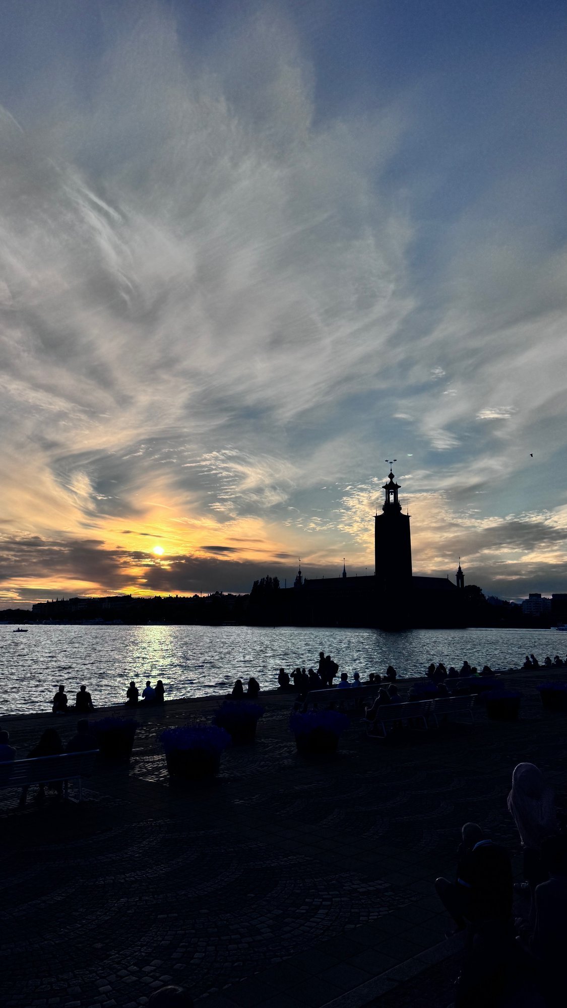 People sit along a stone quay at sunset. Across the water, the tower of Stockholm City Hall stands dark against an orange and gray sky.