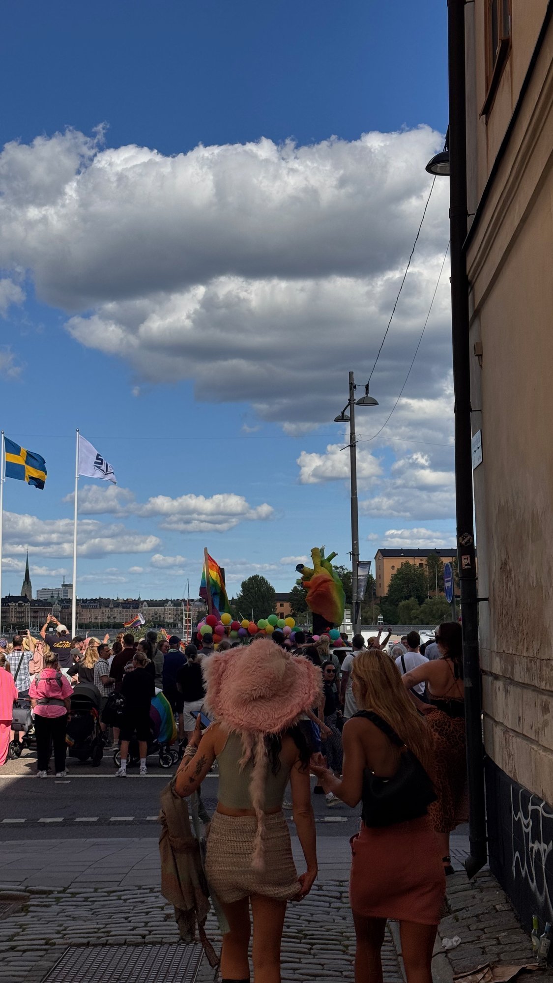 A narrow cobbled lane opens onto the waterfront, where a crowd in rainbow colors watches the Pride parade. A Swedish flag flies on a pole.