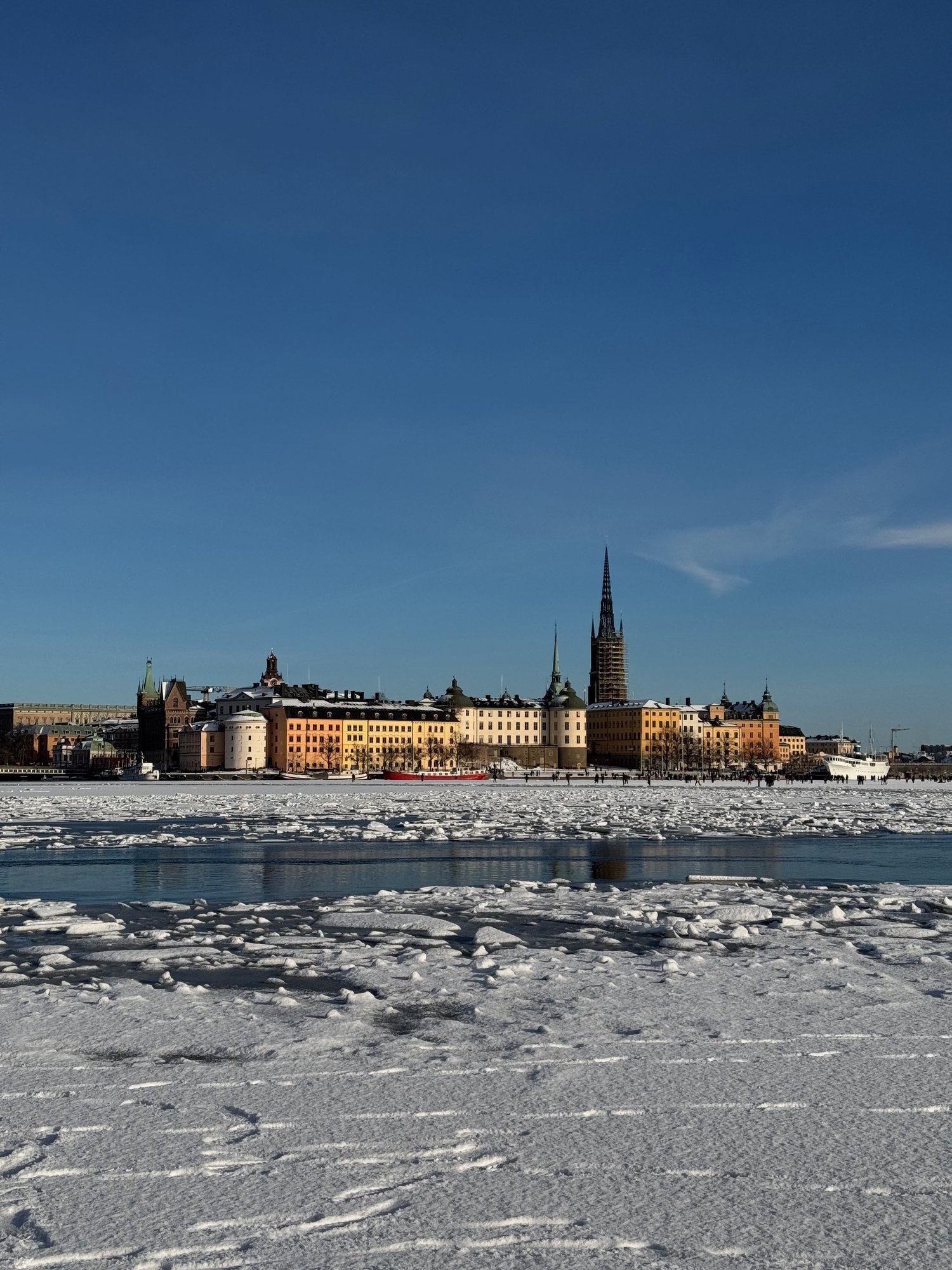 Snow and ice cover Riddarfjärden. Across it stand the old buildings of Riddarholmen and the open iron spire of Riddarholmskyrkan, under a clear blue sky.