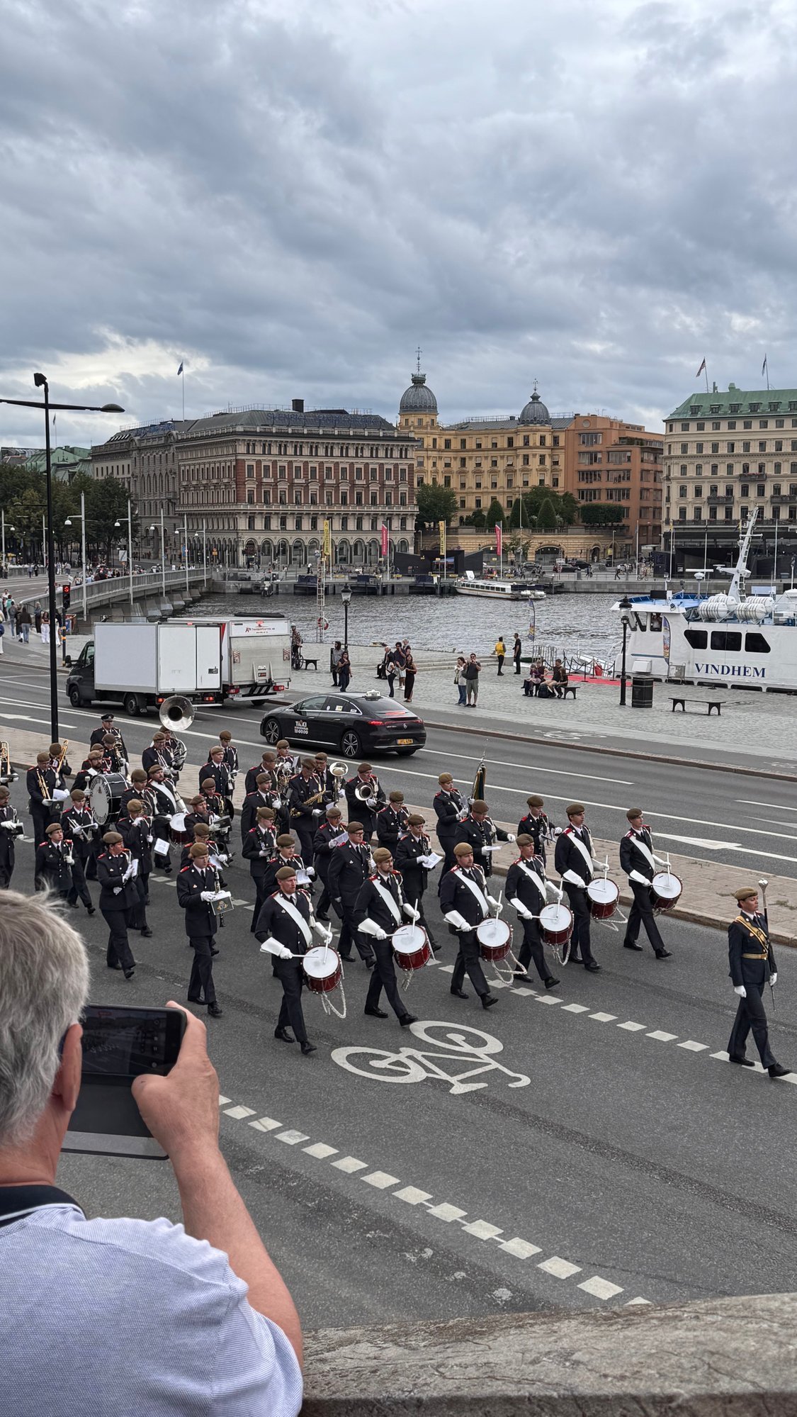 A military band in dark uniforms marches along a street by the water. A man in the front takes a photo with his phone.